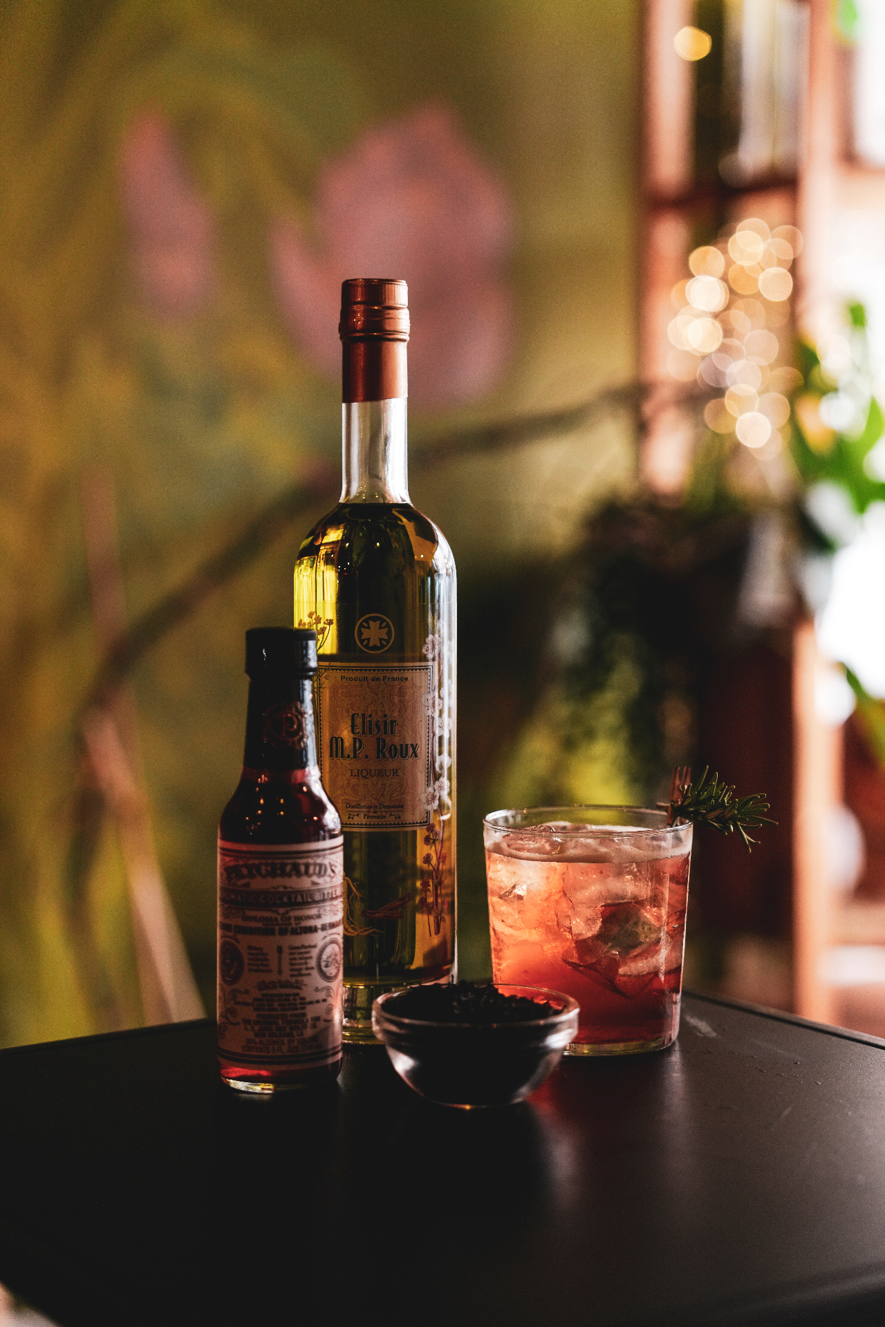 Bouteille d’Elisir M.P. Roux, bitter Bichaud’s et cocktail rouge sur ice servi dans un verre old fashioned avec un brin de romarin, sur une table noire dans une ambiance tamisée de bar à cocktails. Photo Up For It, Murphy VM.