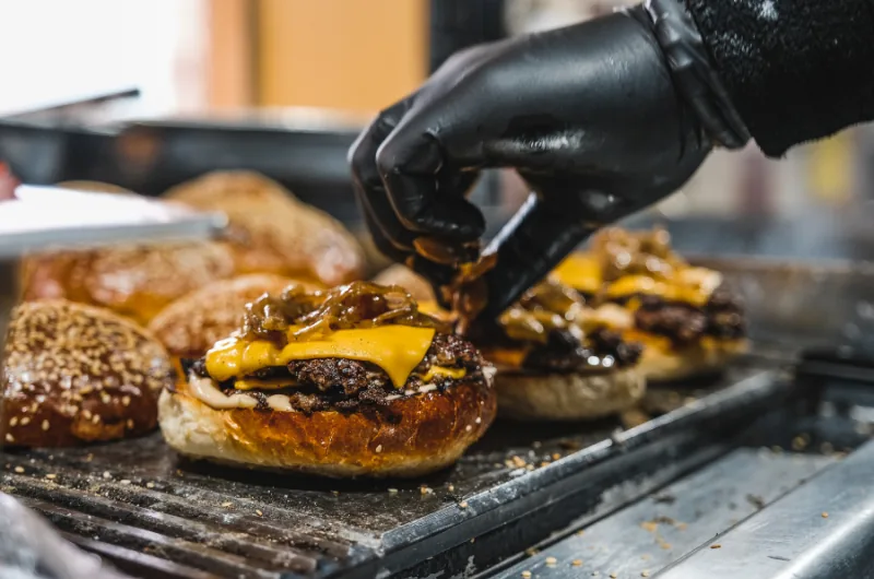Main gantée de noir dressant des smash burgers au cheddar fondu et oignons caramélisés sur une plancha.