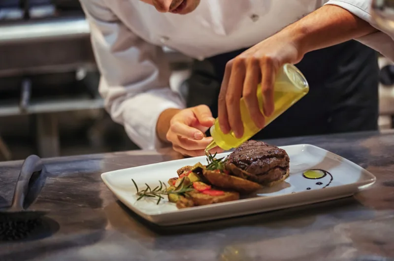 Chef en cuisine versant un filet d'huile d'olive sur une assiette dressée, dans un micro-restaurant.